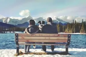 woman, dog and man sitting on bench. mountains and lake surrounding them